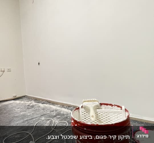 דלי צבע טמבור ומברשת על רקע קיר לבן שנצבע בחדר ריק